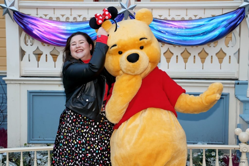 Sam Doyle, dressed in a polkadot skirt, black leather jacket and red turtle neck top, holds Minnie mouse ears above Winnie The Pooh's head as he bashfully stares at the camera.