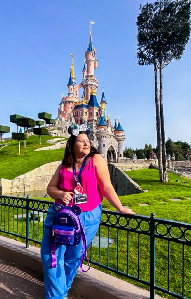 Sam Doyle stood at the garden railing infant of Sleeping Beauty's Castle, Disneyland Paris. She's wearing a bright pink top, blue trousers and carrying a purple 30th anniversary lounge fly bag