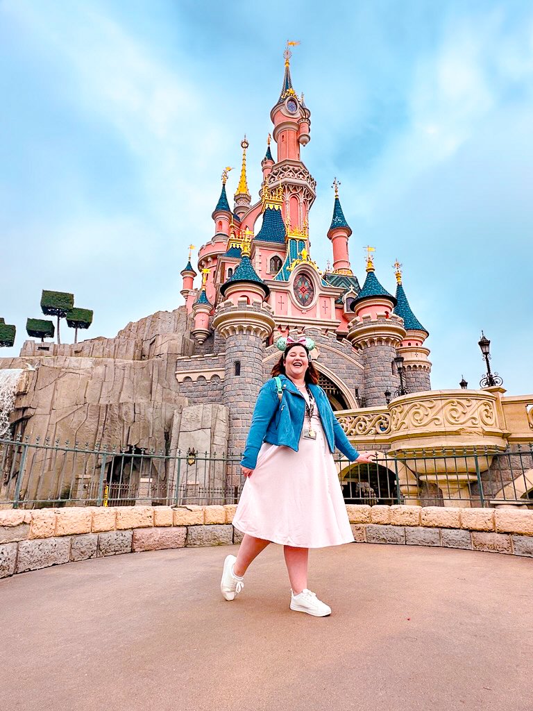 Sam stands infront of the Sleeping Beauty castle in Disneyland Paris and smiles at the camera. She's wearing a pink dress and a blue short jacket.