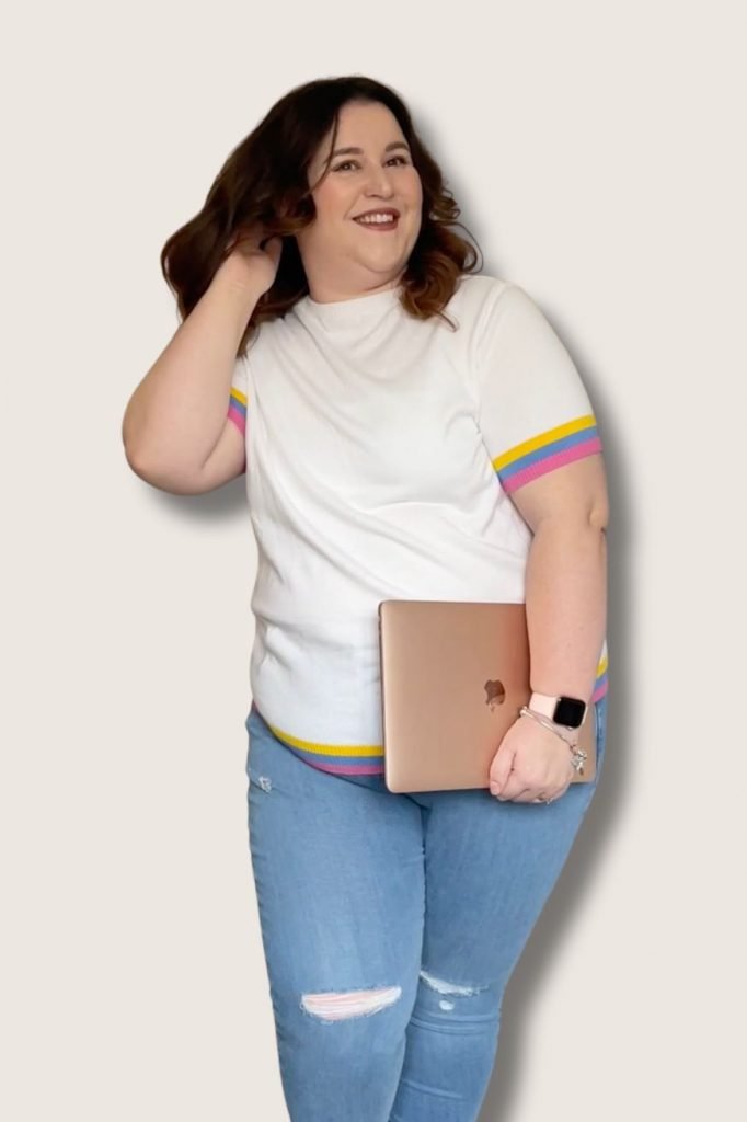 Sam, the create of the Pixie Dust Wanderlust Disneyland Paris Blog, and your Disneyland Paris guide, stands against a beige background. She's wearing blue ripped jeans, a white jumper with a yellow, blue and pink stripe on the sleeve, holding her MacBook and flicking her hair while smiling.